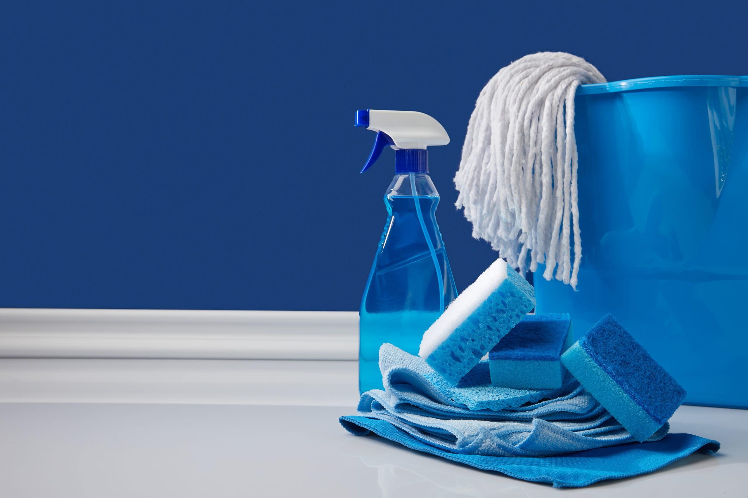 Blue rags, spray bottle, mop and bucket stand next to a darker blue wall.