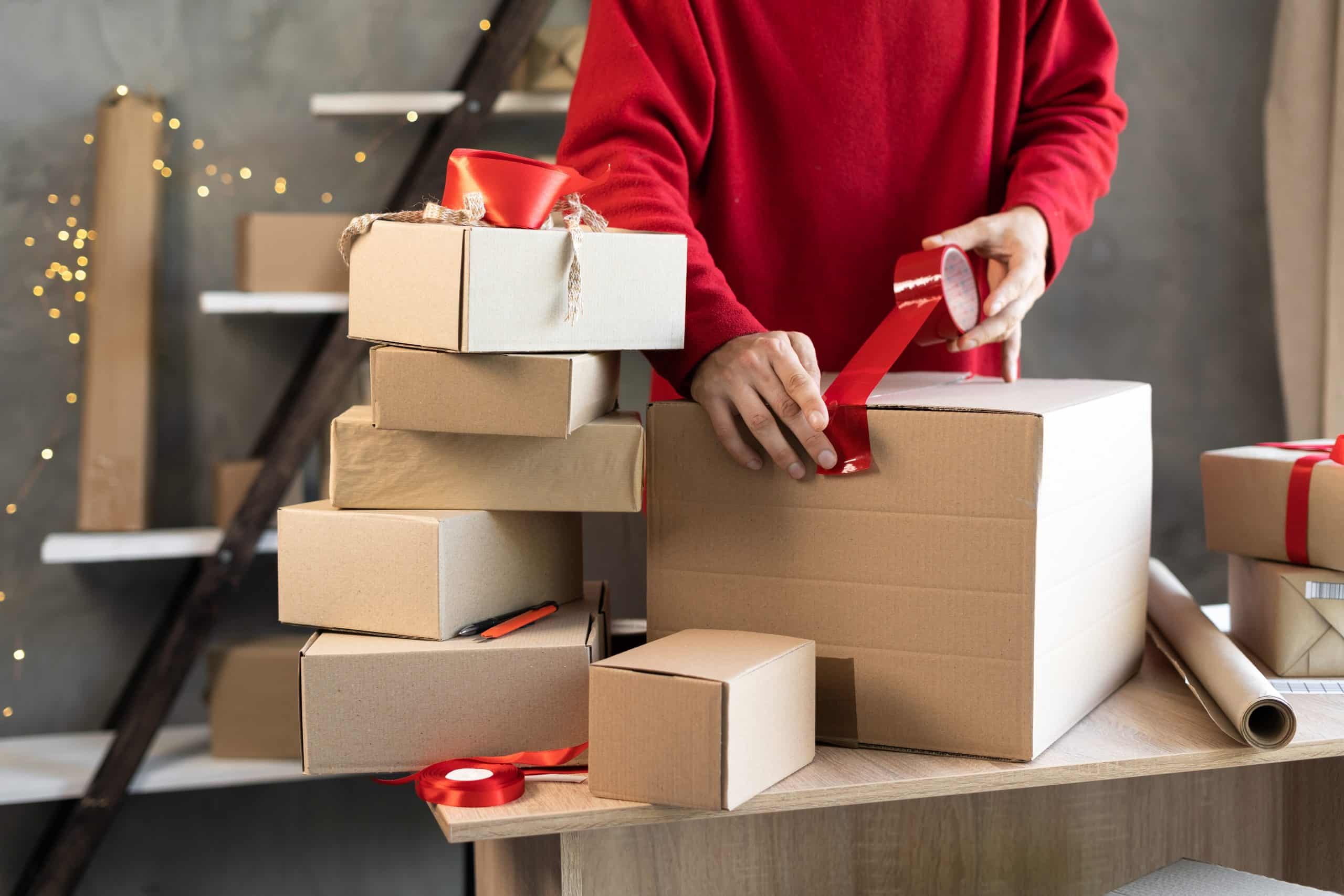 Person surrounded by Christmas packaging boxes and adding red tape to one of them