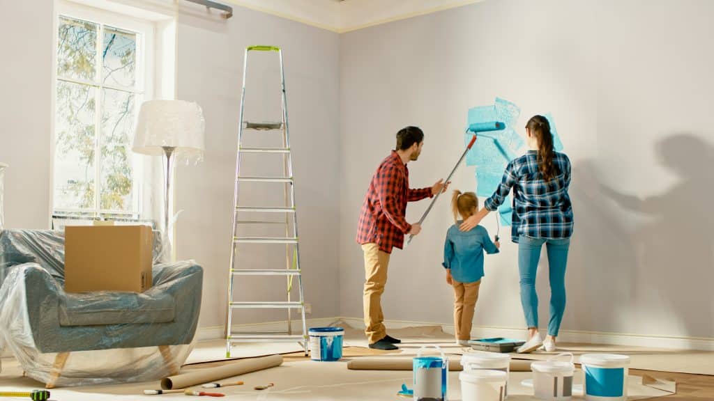 Shows the interior of a living room and a woman, man and child painting the wall in a blue coloured paint with furniture and boxes around them - self storage near Newbury can help during a home renovation