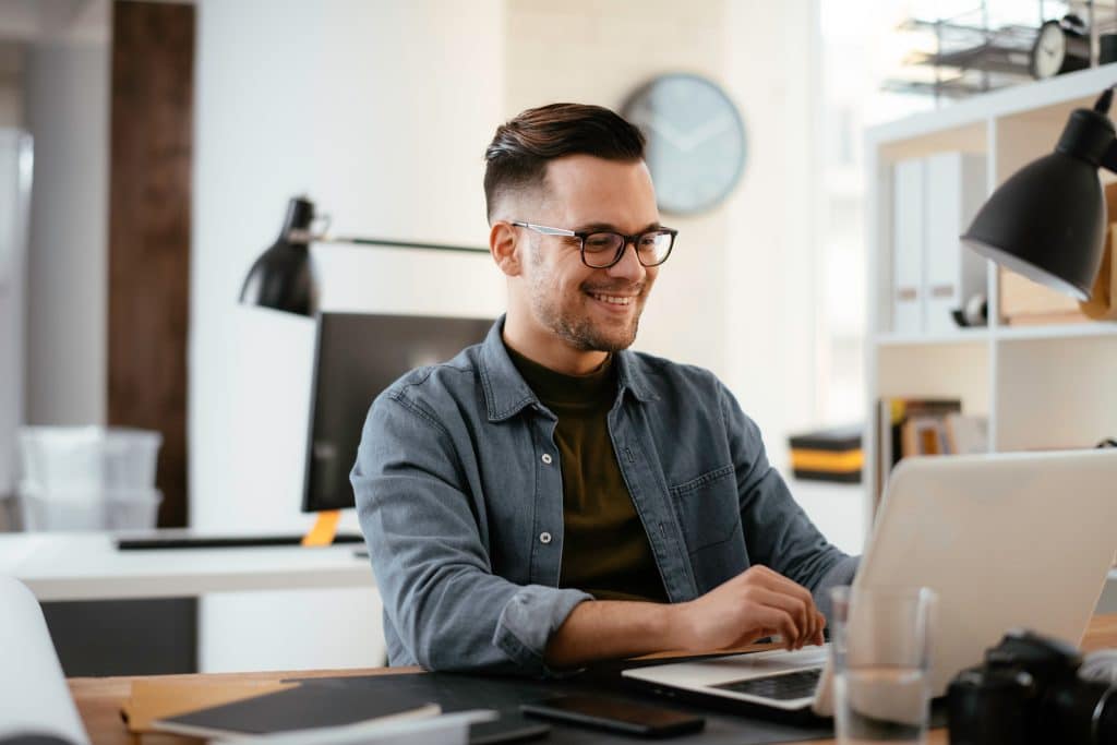 Good mental health in the workplace: How our offices to rent in Chippenham can help. Image shows a man sitting at his desk smiling wearing a denim shirt and dark rimmed glasses.
