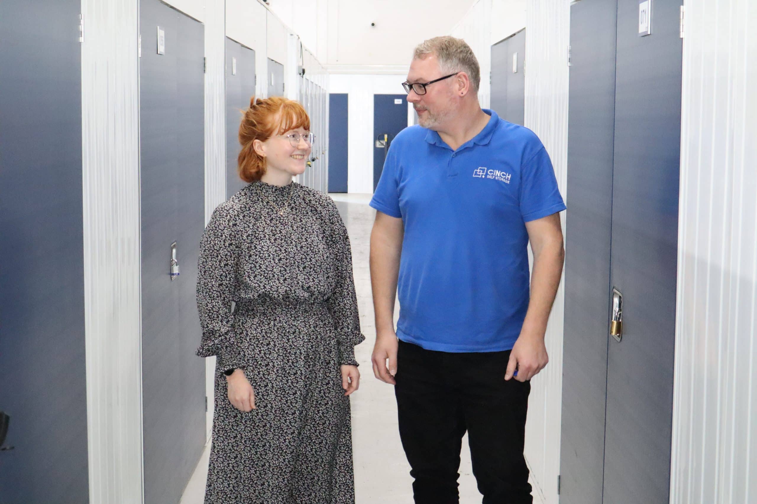 Going home for the summer? Our Brighton storage units will help. Image shows a female student and a member of the Cinch Self Storage team standing in the corridor with storage unit doors on either side of them.