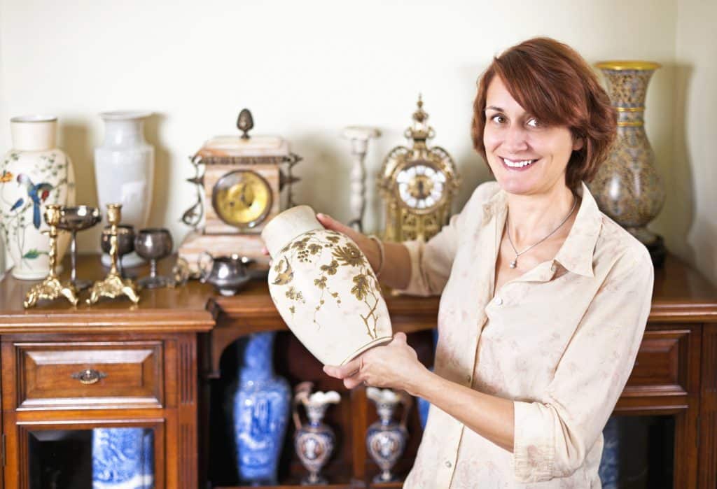 Furniture storage - image shows woman holding an antique vase with display of antiques including clocks and vases on a wooden dresser behind her