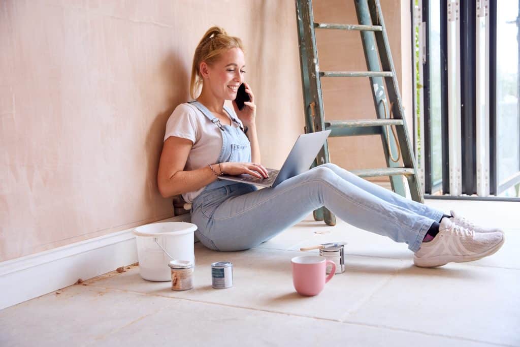 Renovation storage - tips to alleviate stress, image shows woman sitting on the floor in a renovation space with a pink wall holding a phone and a laptop on her knees, surrounded by paint pots and a ladder leaning against the wall