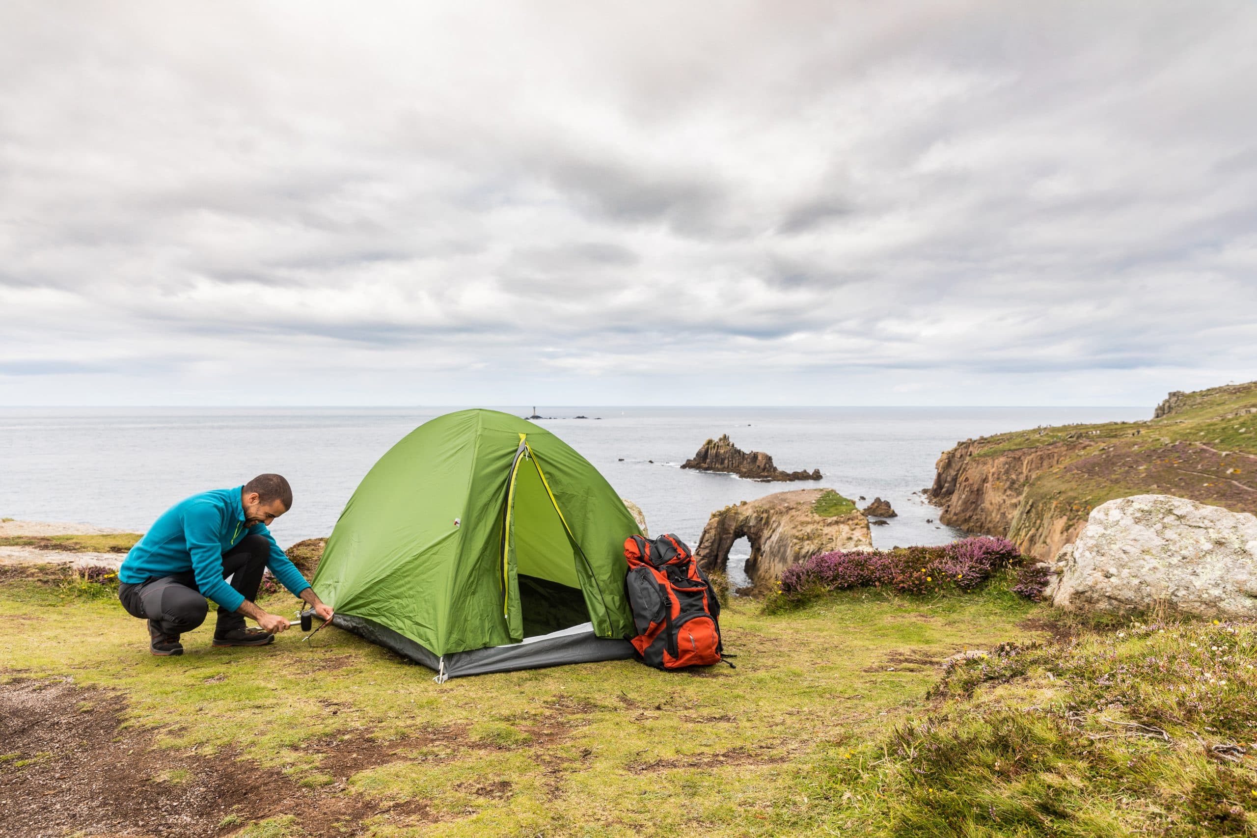 Want to go camping but don’t have the storage? Our Brighton storage will help. Image shows a green tent pitched on a cliff edge overlooking the sea with a man in a blue jumper securing the tent into the ground.