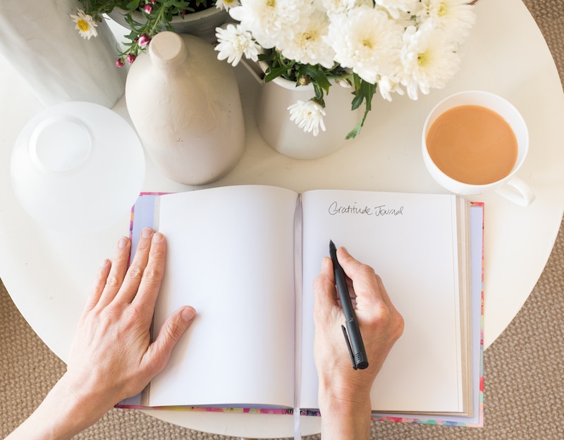 Storage Wandsworth. Image shows an open gratitude journal on a white table with a person's arms holding a pen over the journal. A cup of tea is on the right of the journal with white flowers also on the table.