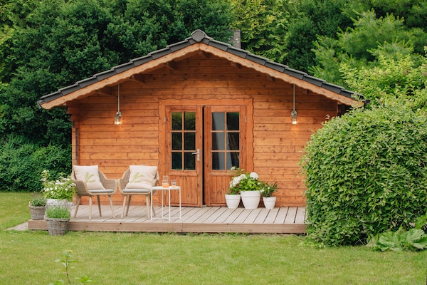 Storage in Chippenham. Image shows the exterior of a summer house situated on a green lawn surrounded by trees. With two chairs, a table and potted plants on the decking. 