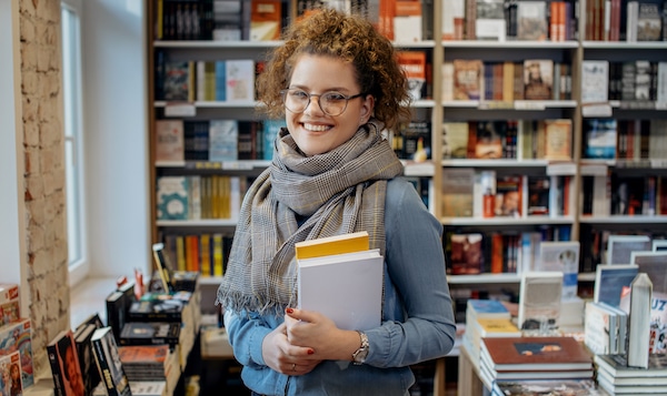 Self-storage Bicester. Image shows a woman with curly hair in a ponytail and glasses standing infront of a bookshelf holding two books - one yellow and one white wearing a grey woollen scarf. 