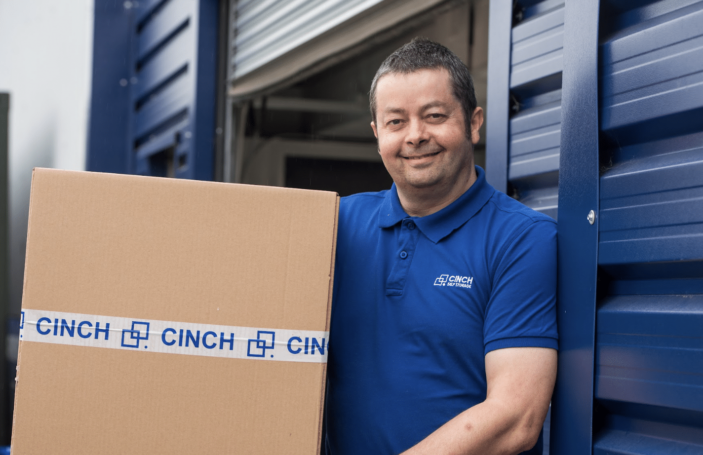 benefits_of_short_term_storage. Image shows a Cinch self storage employee wearing a blue branded top and holding a Cinch self storage cardboard box in front of shutter doors.