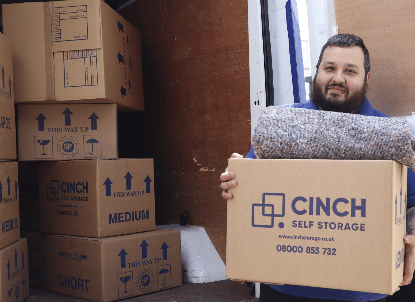 moving_to_newbury. Image shows cinch self storage employee holding a cinch cardboard box with a dust blanked on top smiling with an open van filled with cinch self storage boxes.