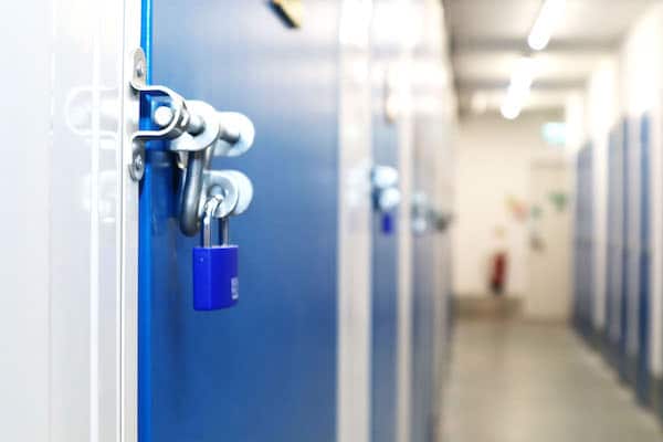 Secure self storage. Image shows a close up of a blue padlock on a blue door of a self storage unit, with other storage units along the corridor.