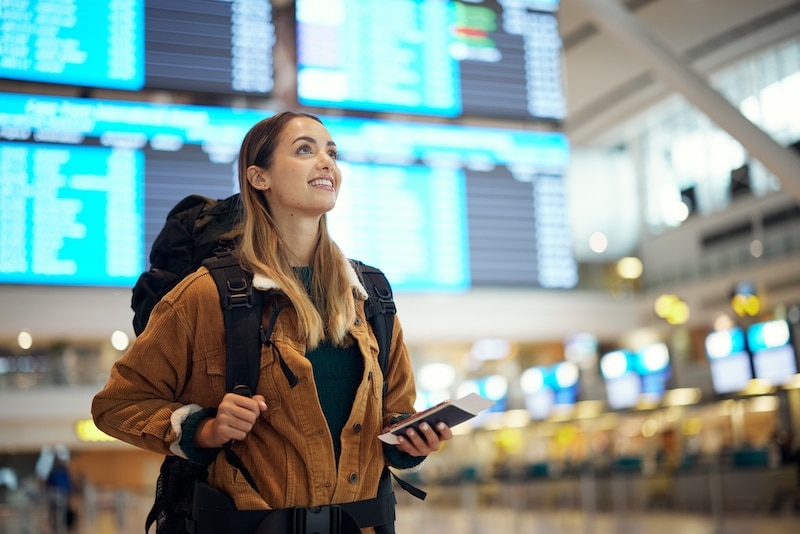 Storage for travellers. Image shows a young woman standing infont of airport screens holding her phone, with a backpack on her back looking up and smiling.