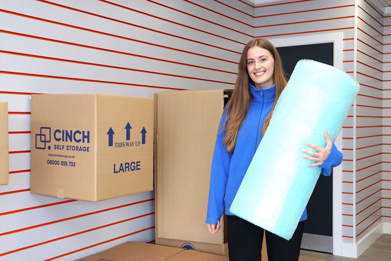 Essential items for efficient self storage packing. Image shows a woman in a blue Cinch Self Storage jumper holding blue bubble wrap and standing in front of a cardboard box display. 