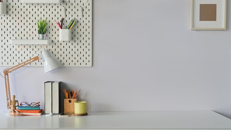 Office to rent in Brentwood. Image shows a desk in an office, with a light grey wall with a pin board with pen pot, white lamp and books with a yellow pencil pot on the desk.