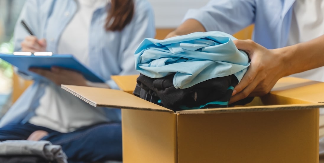 9 Essential tips for storing clothes long-term. Image shows people putting clothes in a cardboard box with someone holding a blue clipboard in the background.