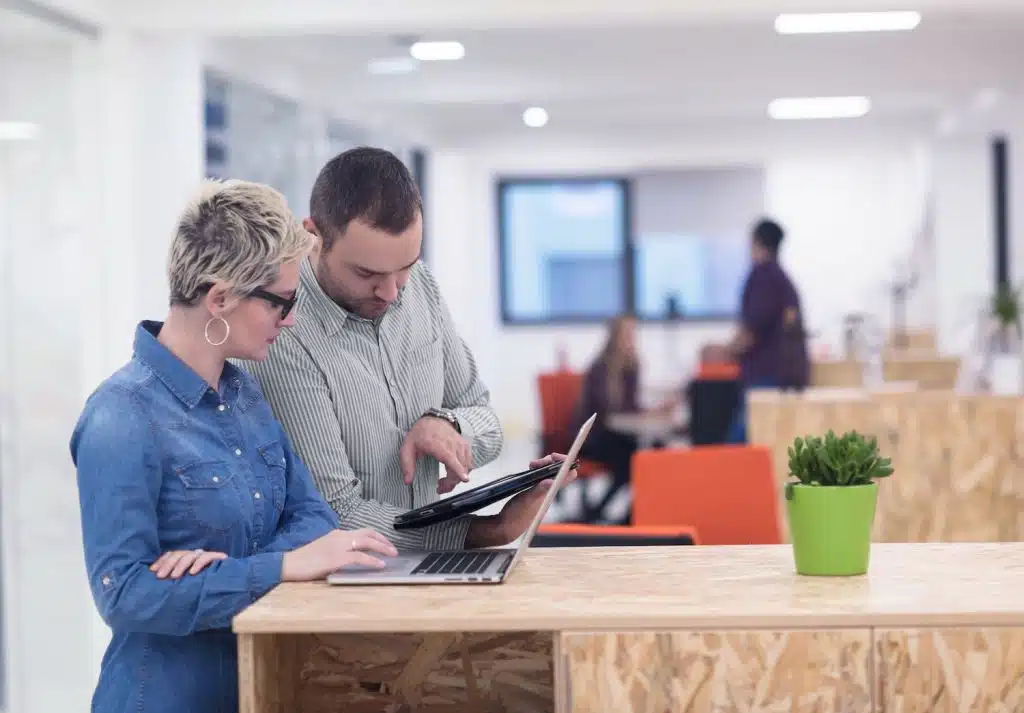 Office to rent in Leighton Buzzard. Image shows a man and a woman pointing on a laptop on a chipboard desk with a white office behind with windows in the back.