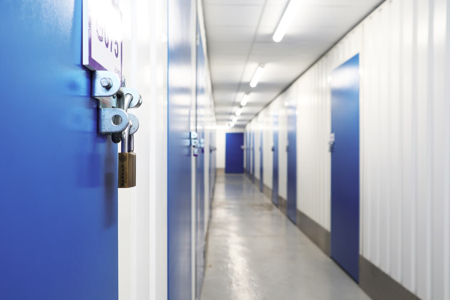 Storage unit in Mitcham. Image shows a close up view of a padlock on a storage unit blue door. With a corridor of storage units beyond.