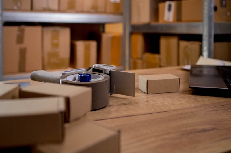 6 Benefits of business storage in Letchworth. Image shows shelving behind a desk filled with cardboard boxes and a tape dispenser and more cardboard box parcels on top of the desk.