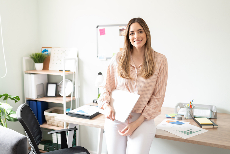 9 Benefits of serviced offices for modern businesses. Image shows a woman holding a laptop in a light pink top, sitting on a light wooden desk with shelving to the side of her and a noticeboard behind her