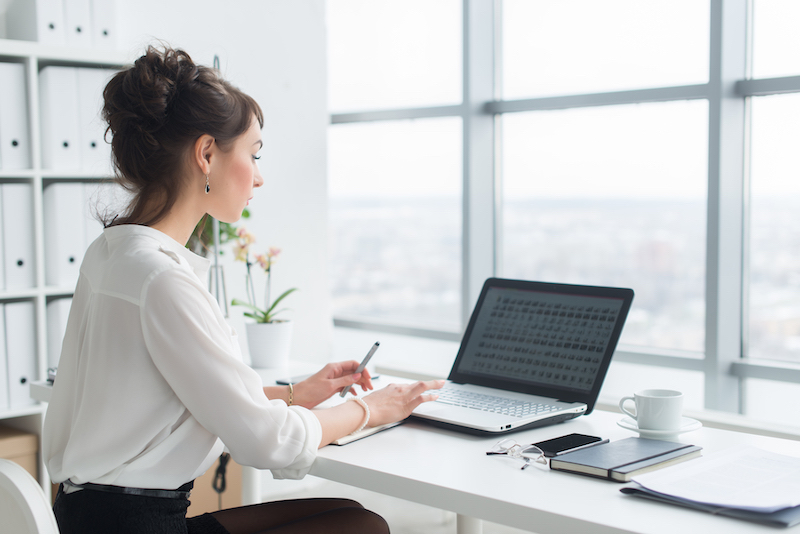Exploring types of offices from serviced to virtual workspaces. Image shows a businesswoman wearing a white shirt sitting at her desk, typing on her laptop. With a light and airy interior with large windows