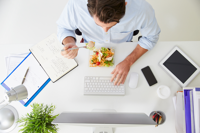 How much office space do you need? Office space per person. Image shows an overhead view of a businessman working at a computer in an office eating a salad in a blue shirt