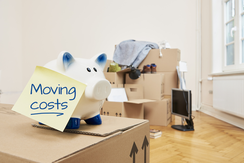 4 Unexpected Moving House Costs. Image shows a piggy bank on top of a cardboard box, which has a yellow piece of paper stuck to it.