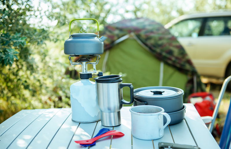 7 Camping equipment storage tips. Image shows a green tent in the background and silver flasks, a mug and crockery on a camping table in the foreground.