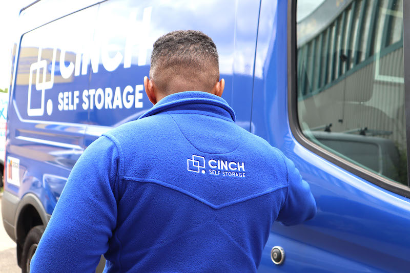 Self storage in Dunstable. Image shows a Cinch Self Storage employee with their back to camera wearing a blue Cinch fleece and standing in front of the blue Cinch Self Storage van.