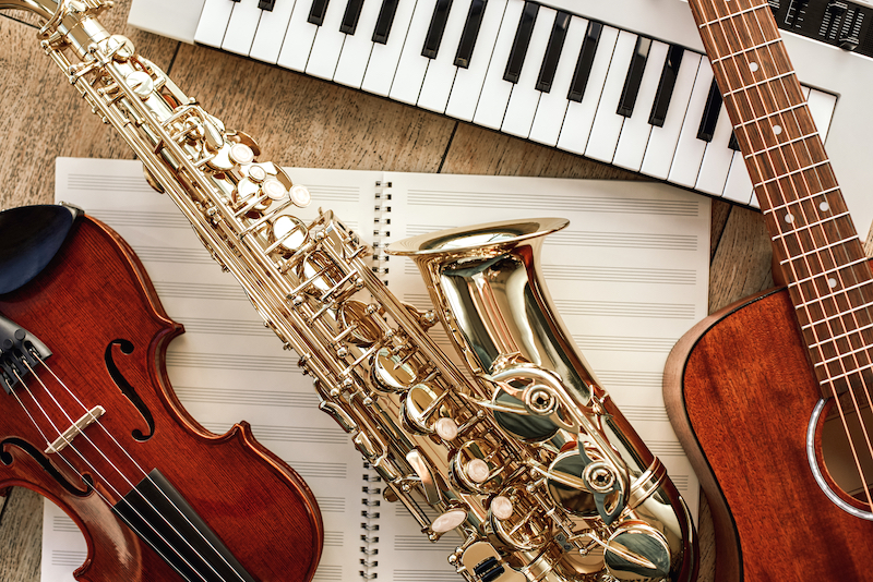 6 Essential tips for storing musical instruments safely. Power of music. Image shows top view of musical instruments including a synthesizer, guitar, saxophone and violin lying on the sheets for music notes on a wooden floor.
