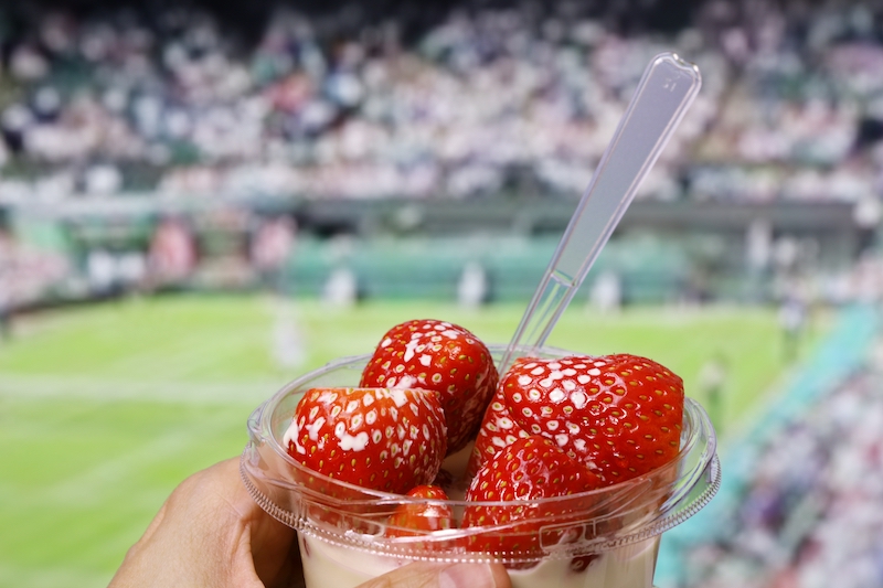 The ultimate guide to things to do in Wimbledon. Image shows a hand holding strawberries and cream in a small bowl, overlooking a tennis court.