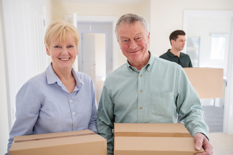 House move storage Sittingbourne. Image shows a senior Couple Downsizing In Retirement Carrying Boxes Into A New Home On Moving Day With Removal Man Helping