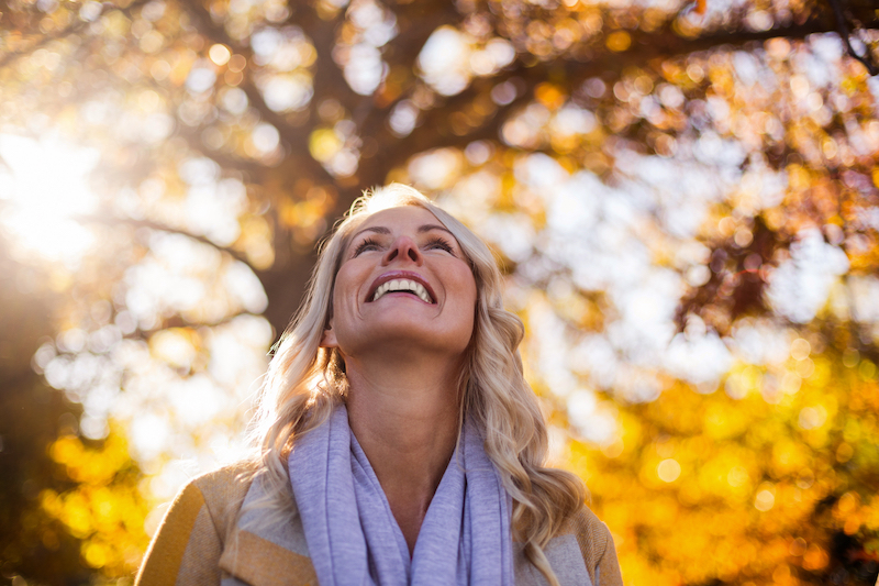 Image shows a smiling woman looking up against trees at park during autumn