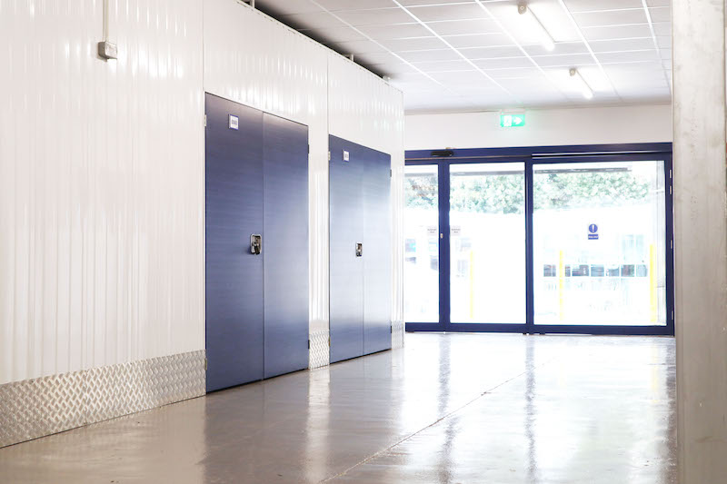 Storage rooms Earlsfield. Image shows blue doors of storage rooms and a bright and light corridor with blue framed glass sliding doors at the end