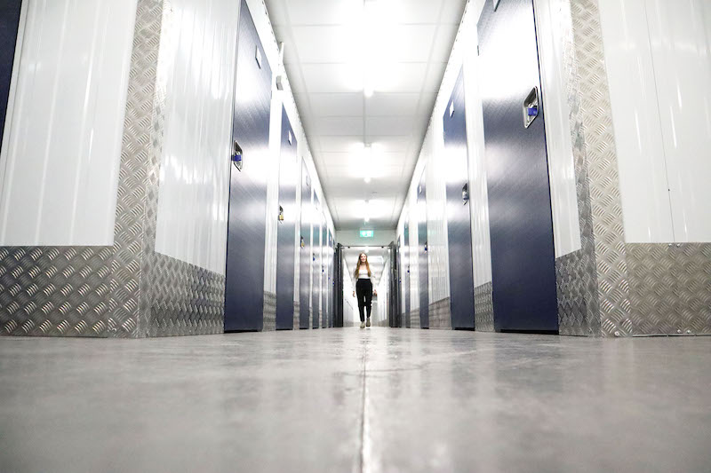 Storage space Seaford. Image shows a woman waling down the corridor of a storage unit facility, with blue storage unit doors on either side. 