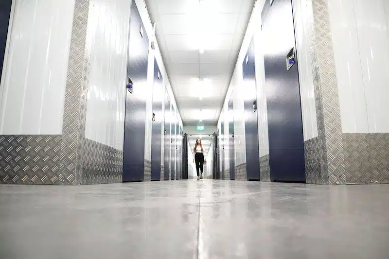 Storage space Seaford. Image shows a woman waling down the corridor of a storage unit facility, with blue storage unit doors on either side.
