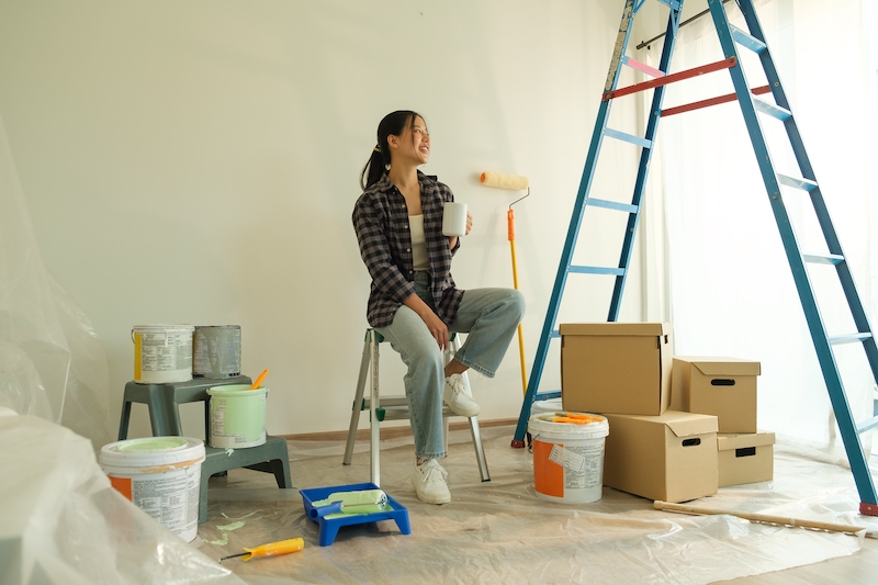 Bedford self storage. Image shows a woman taking a break during a home renovation project, sitting on a step ladder, surrounded by painting supplies.