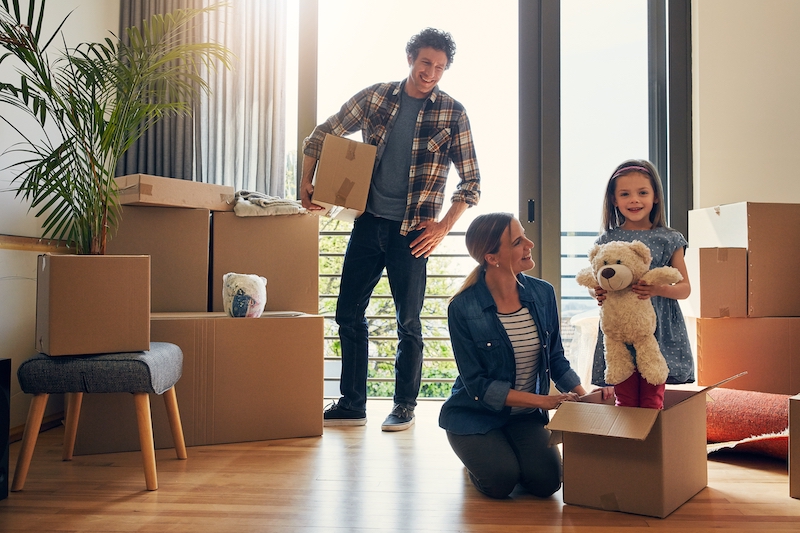 House move storage Watford. Image shows a family moving house standing and laughing with cardboard boxes and a little girl holding a teddy.