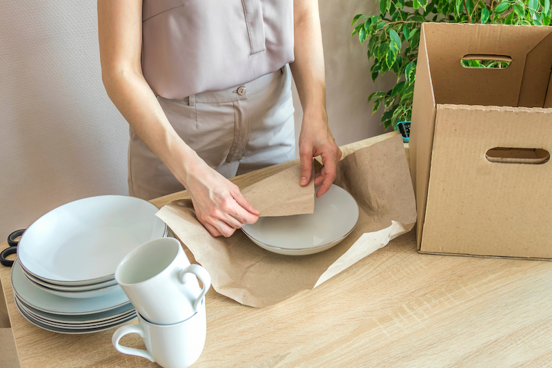 How to pack plates for moving. Images shows a woman wrapping plates in tissue paper ready to pack into cardboard boxes.