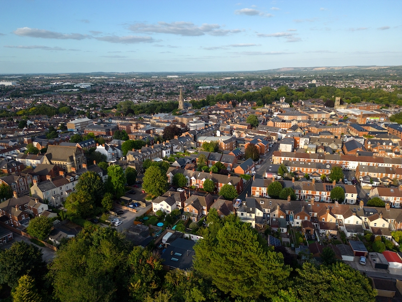 Is Swindon a nice place to live? Image shows a Panorama of Swindon old town in Wiltshire, UK. Aerial view of residential houses with church and landscape at sunset. Above green tree canopy in the summer.