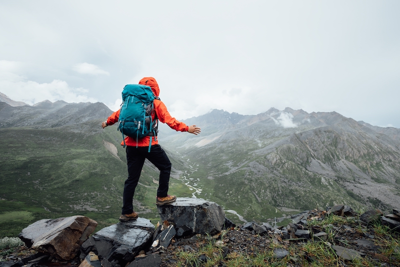 Storage Mitcham. Image shows a person in an orange jacket with green backpack standing on the top of a mountain having hiked up there.