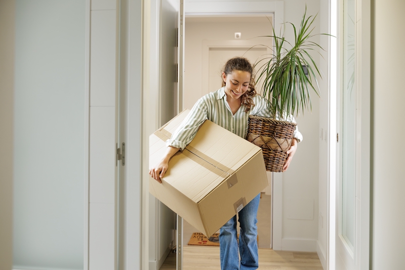 5 Tips for moving out of your parents house for the first time. Image shows a young woman carrying a moving box and a plant.