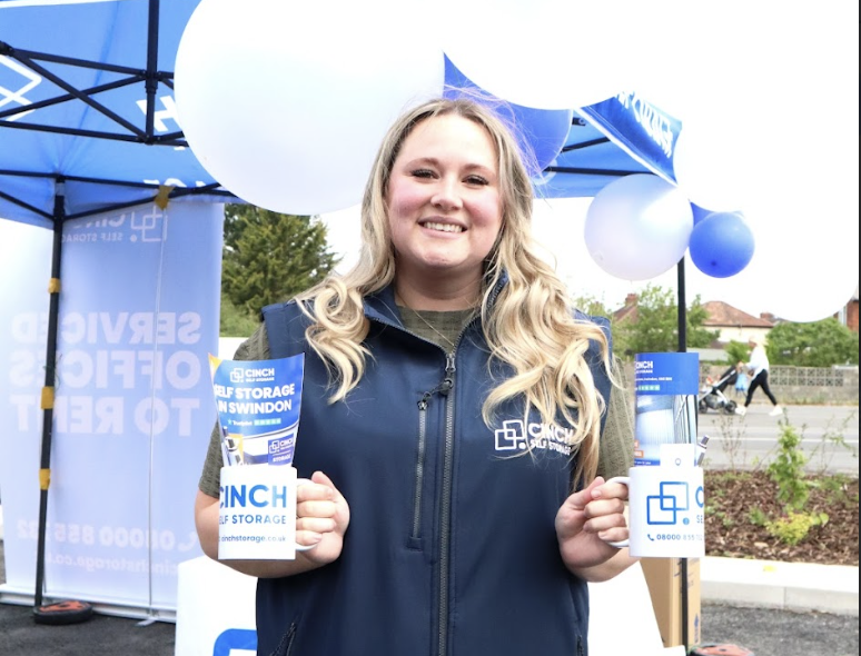Business storage in Wiltshire. Image shows a Cinch Self Storage Employee standing in front of blue and white balloons holding Cinch Storage branded mugs.