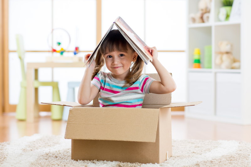 Kids book storage. Image shows a girl sitting in a cardboard box holding a book on her head.