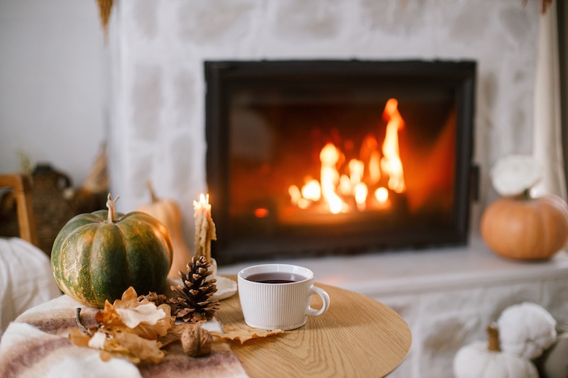Autumn ready. Image shows a warm cup of tea, pumpkin, autumn leaves on wooden table on background of burning fireplace.