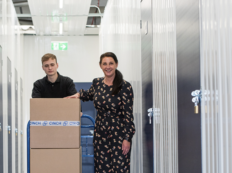 Bicester short term storage. Image shows a woman walking with a Cinch storage employee down the corridor with a trolley piled with boxes.