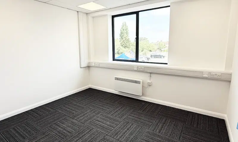 Bicester office to let. Image shows an empty office space with large window, white walls and dark carpet.