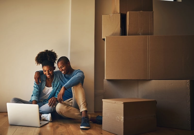 House move storage Paulton. Image shows a couple sitting on the floor surrounded by cardboard boxes moving house and looking at details on their laptop.