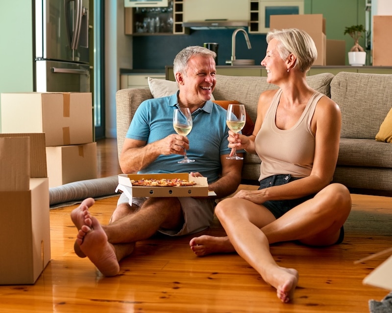 Storage rooms Newcastle. Image shows a mature couple celebrating in their new home on moving day eating pizza and drinking wine on the floor of their new apartment.