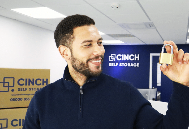 Storage unit Paulton. Image shows a man holding and looking at a gold padlock with Cinch Self Storage cardboard boxes and logo behind him.