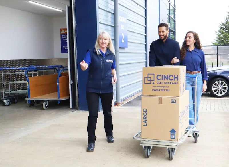 Self storage in Bedford the complete local guide. Image shows a couple being shown into the storage facility by a Cinch Self Storage employee. The couple have a trolley loaded with moving boxes.