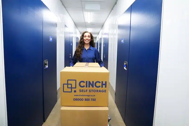 Self storage for newcastle gateshead and tyneside. Image shows a woman walking towards the camera in a corridor of self storage units with a trolley filled with Cinch Self Storage boxes.